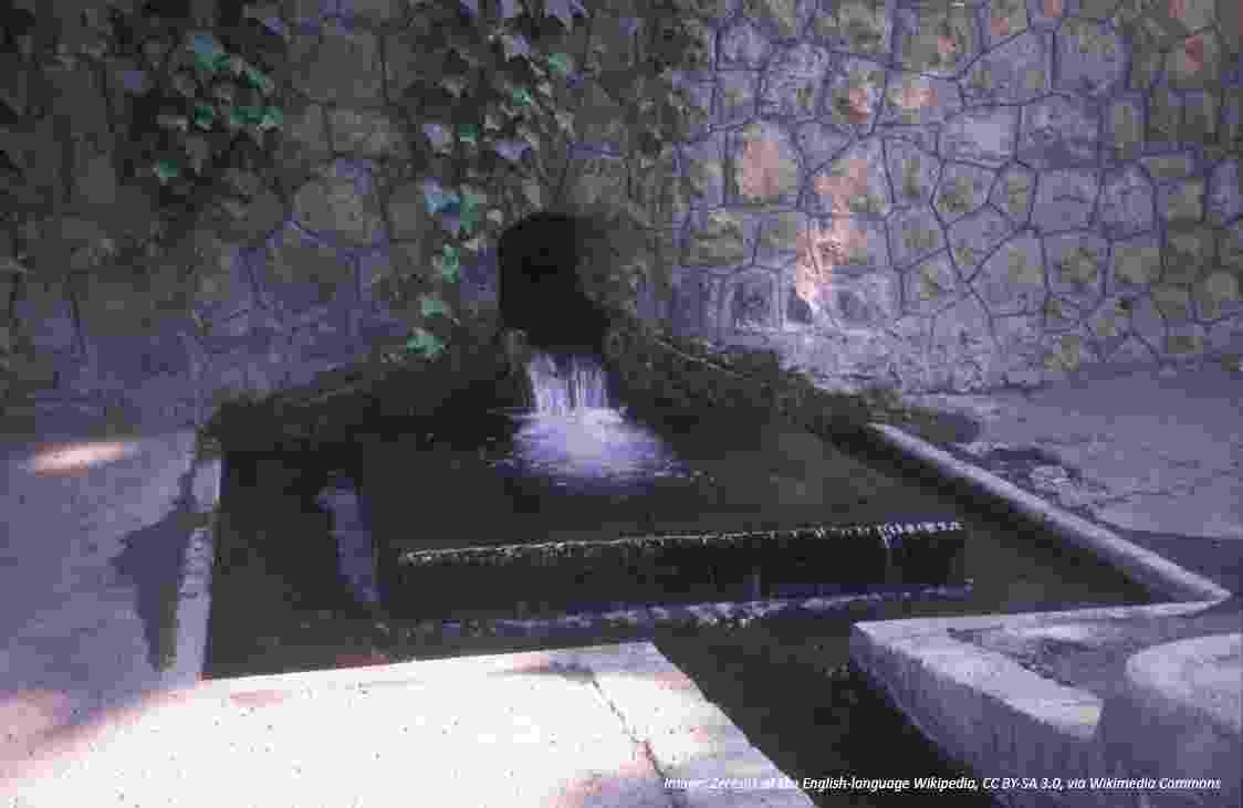 A water spring bubbles from a stone wall into a square fountain well and channel. 